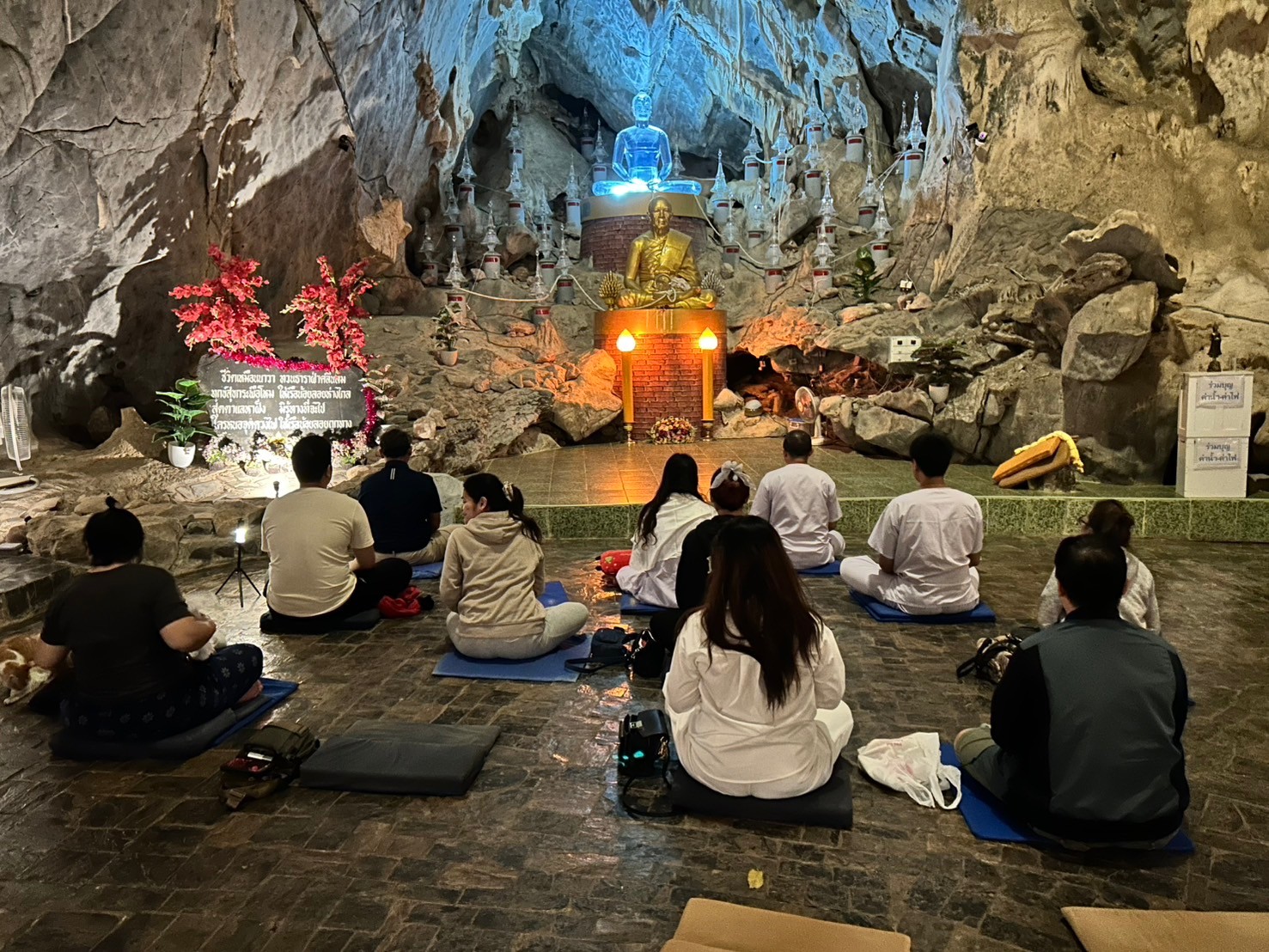 ผู้ปฏิบัติธรรมนั่งสมาธิเบื้องหน้าพระประธานสีทองและพระแก้วสีฟ้าภายในถ้ำ